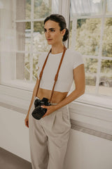 Mujer sosteniendo una cámara con una correa, de pie en una habitación con grandes ventanas.