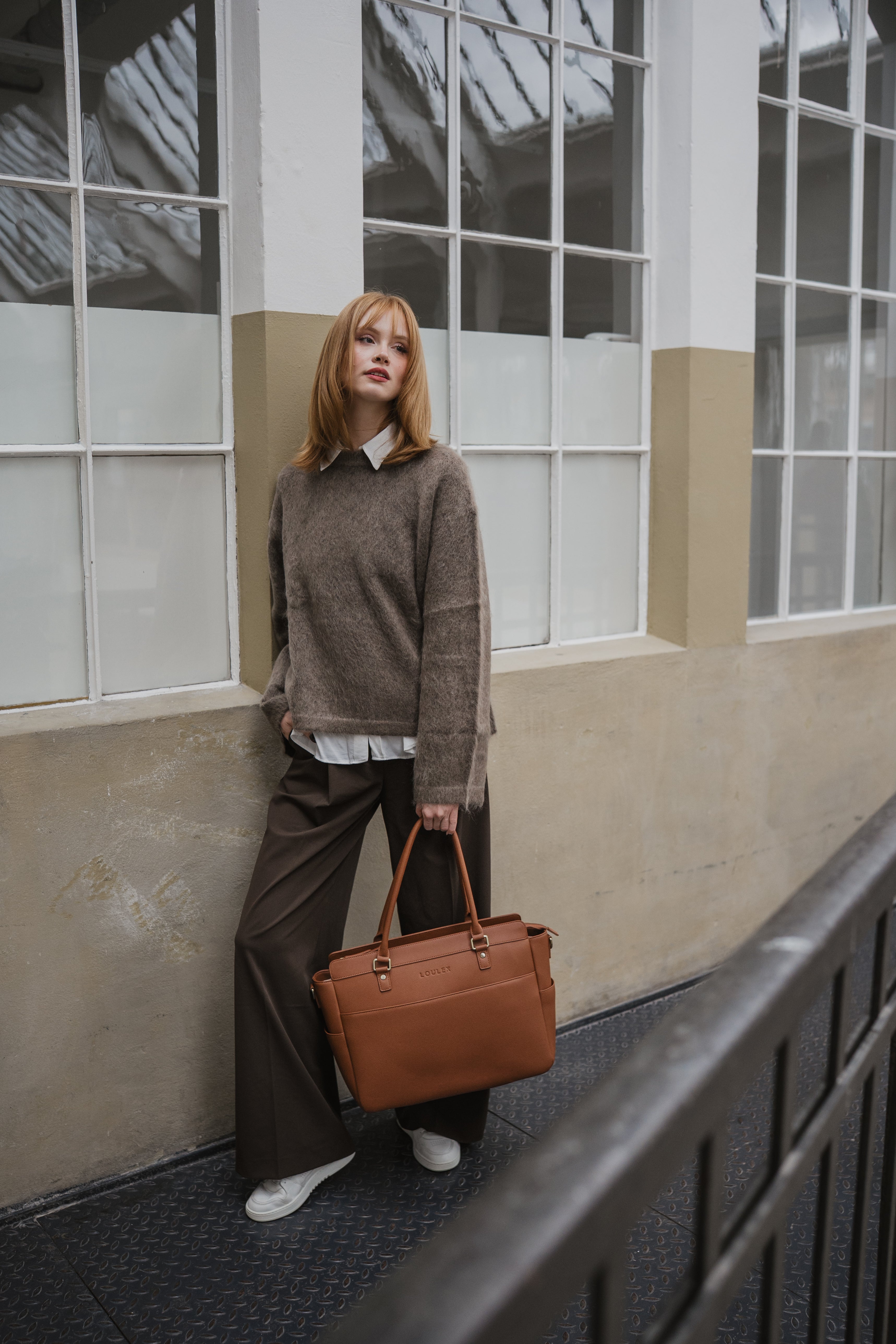 Woman holding a brown leather bag against a building with large windows.