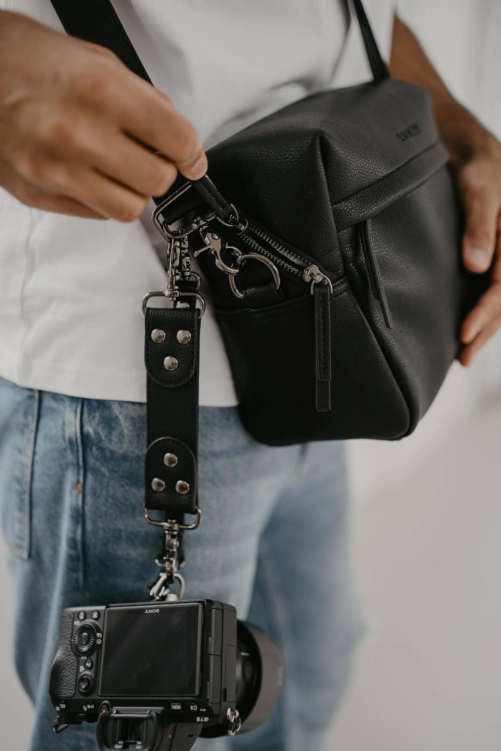 Persona con una bolsa negra para cámara con una cámara acoplada, vestida con una camisa blanca y vaqueros azules.