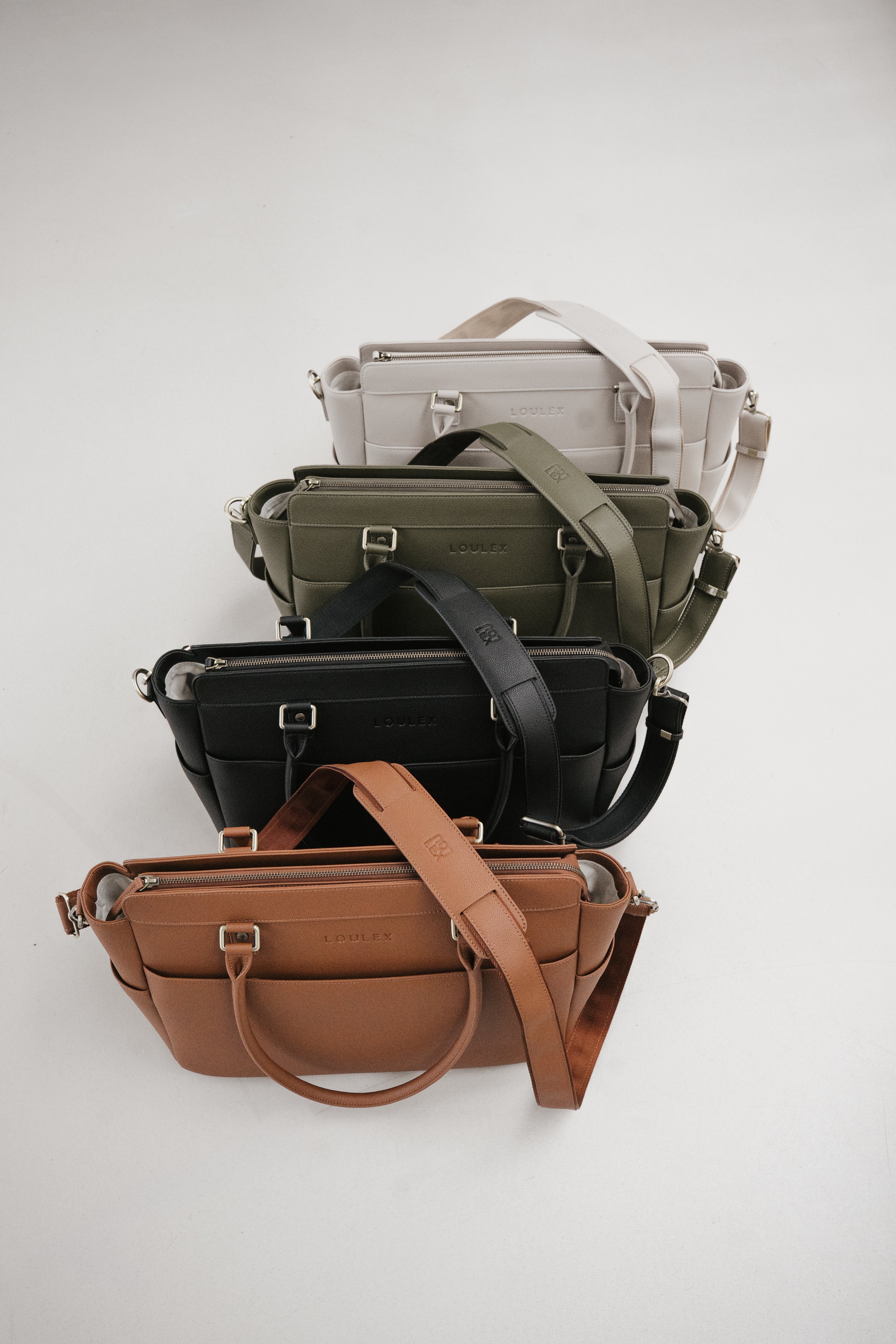 Stack of various bags including a brown leather bag, black leather bag, green leather bag, and gray leather bag on a white background.
