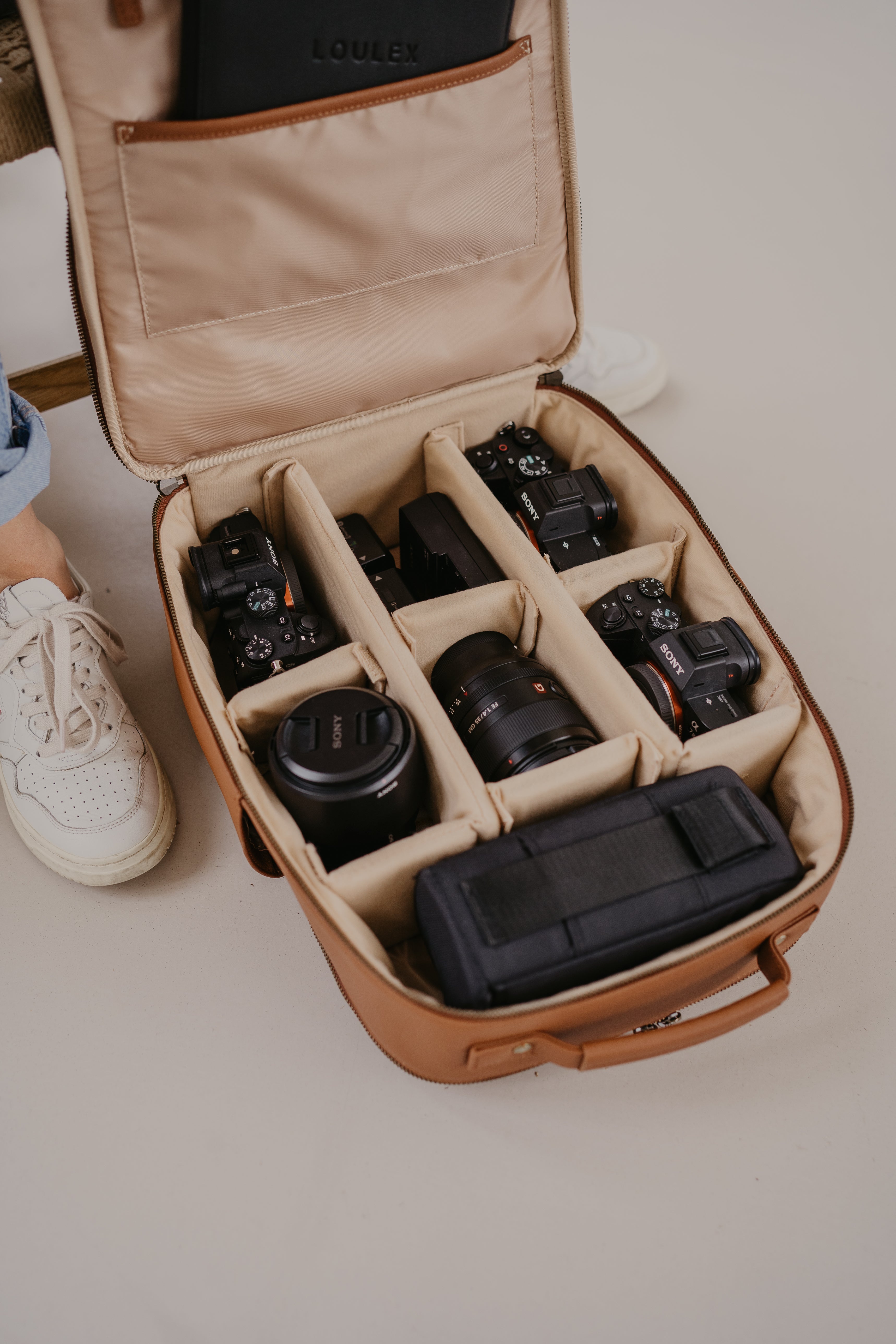 A woman in a grey sweater dress wearing a loulex black leather camera backpack while taking a photo in a minimalist industrial studio.