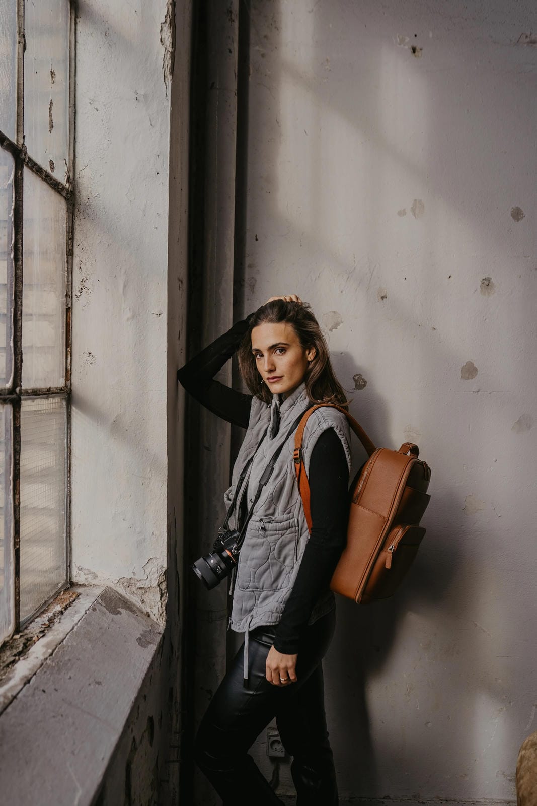 Person leaning against a wall with a camera and backpack in an industrial setting