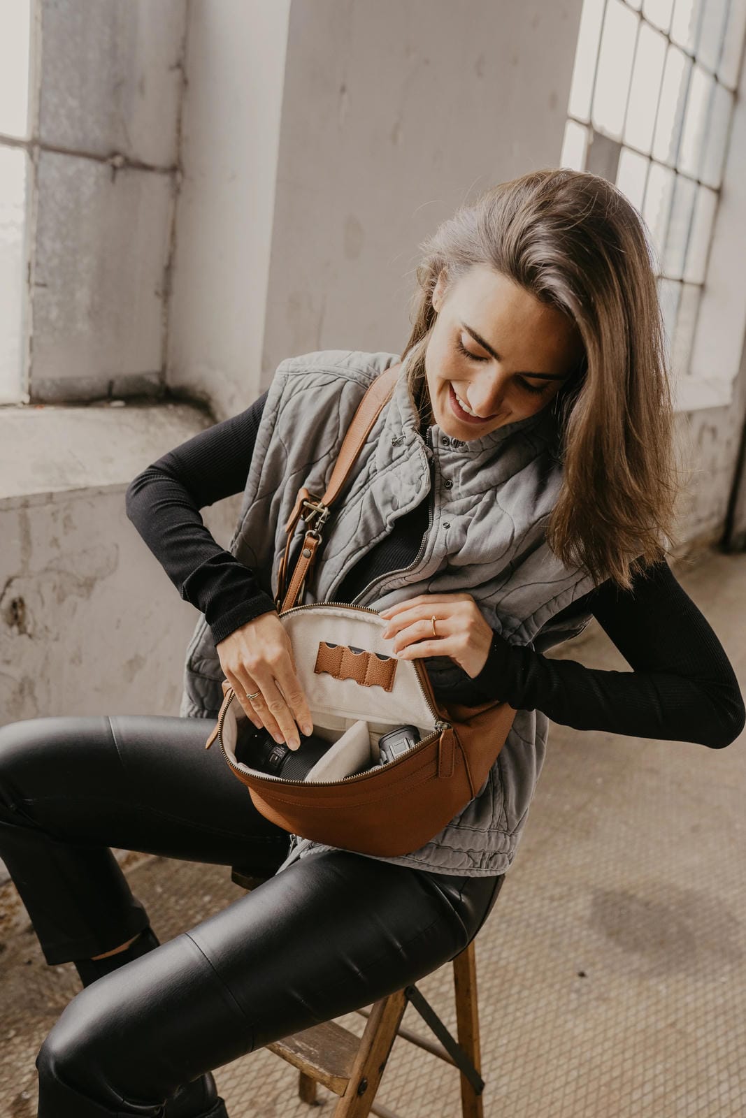 Woman sitting on a stool holding a brown leather bag in an indoor setting.