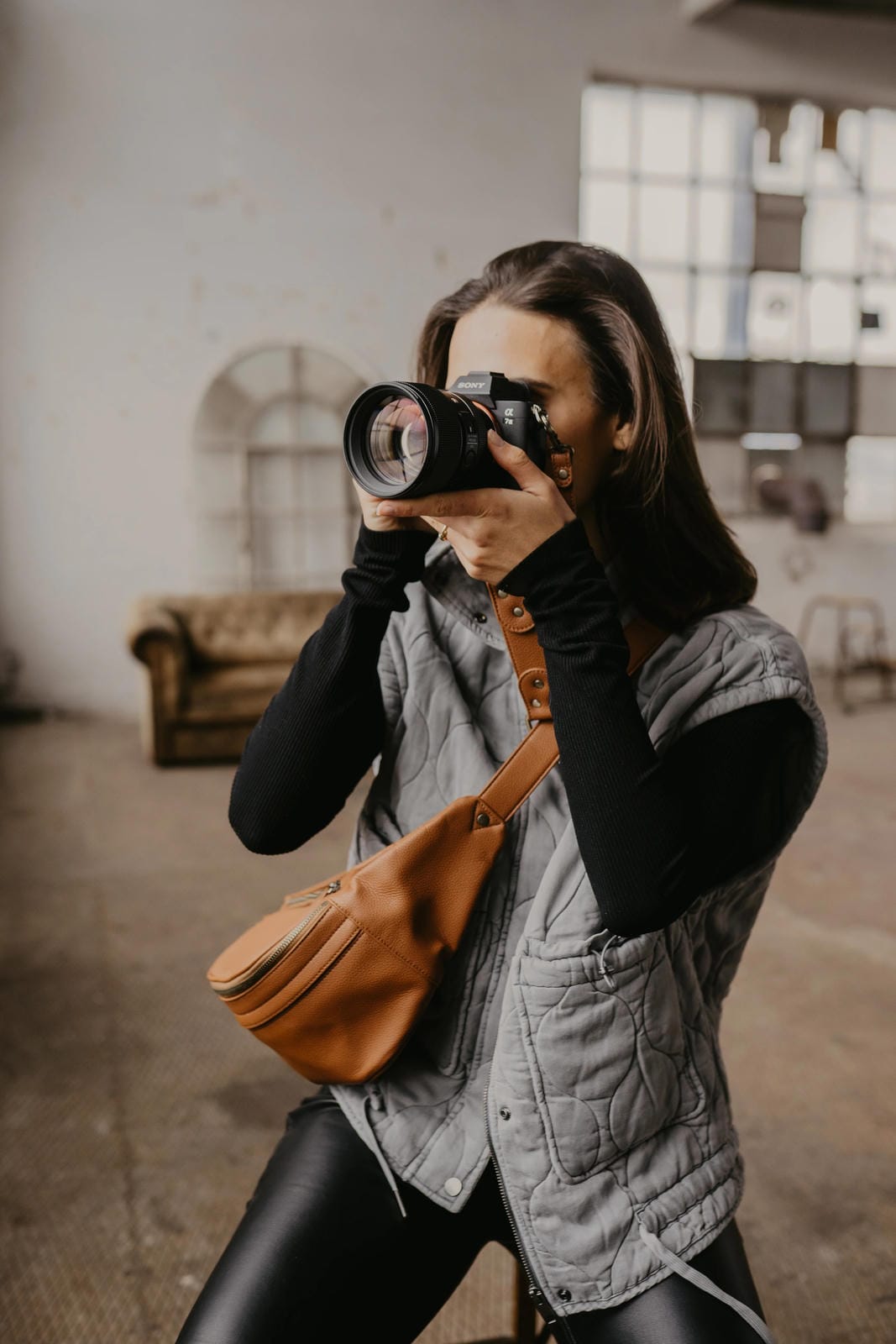 Person holding a camera in an indoor setting with a brown bag over one shoulder.