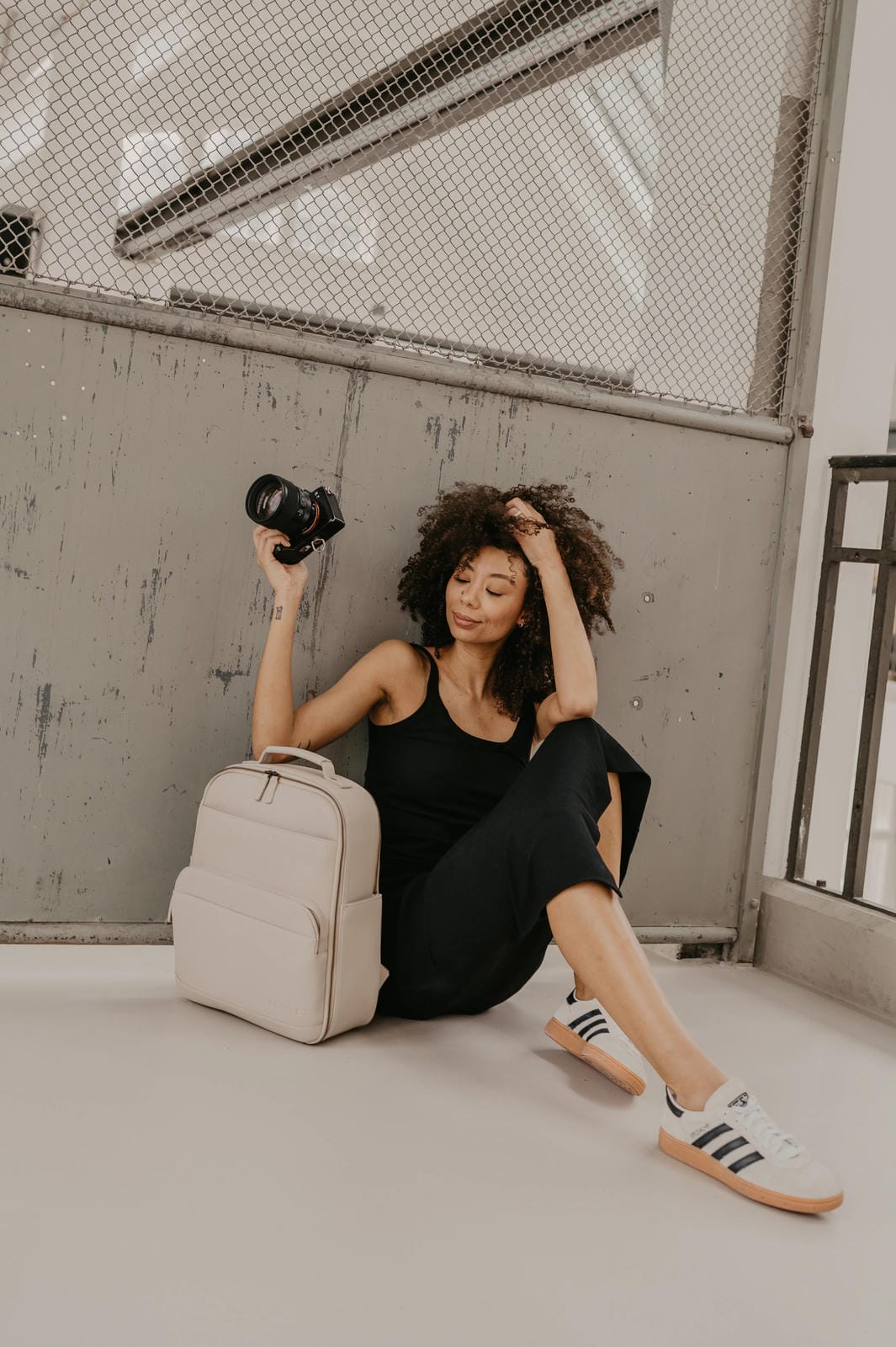 Woman sitting on the ground with a beige backpack and camera, wearing black pants and white sneakers.