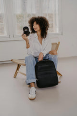 Donna con una macchina fotografica e uno zaino nero in una stanza minimalista.