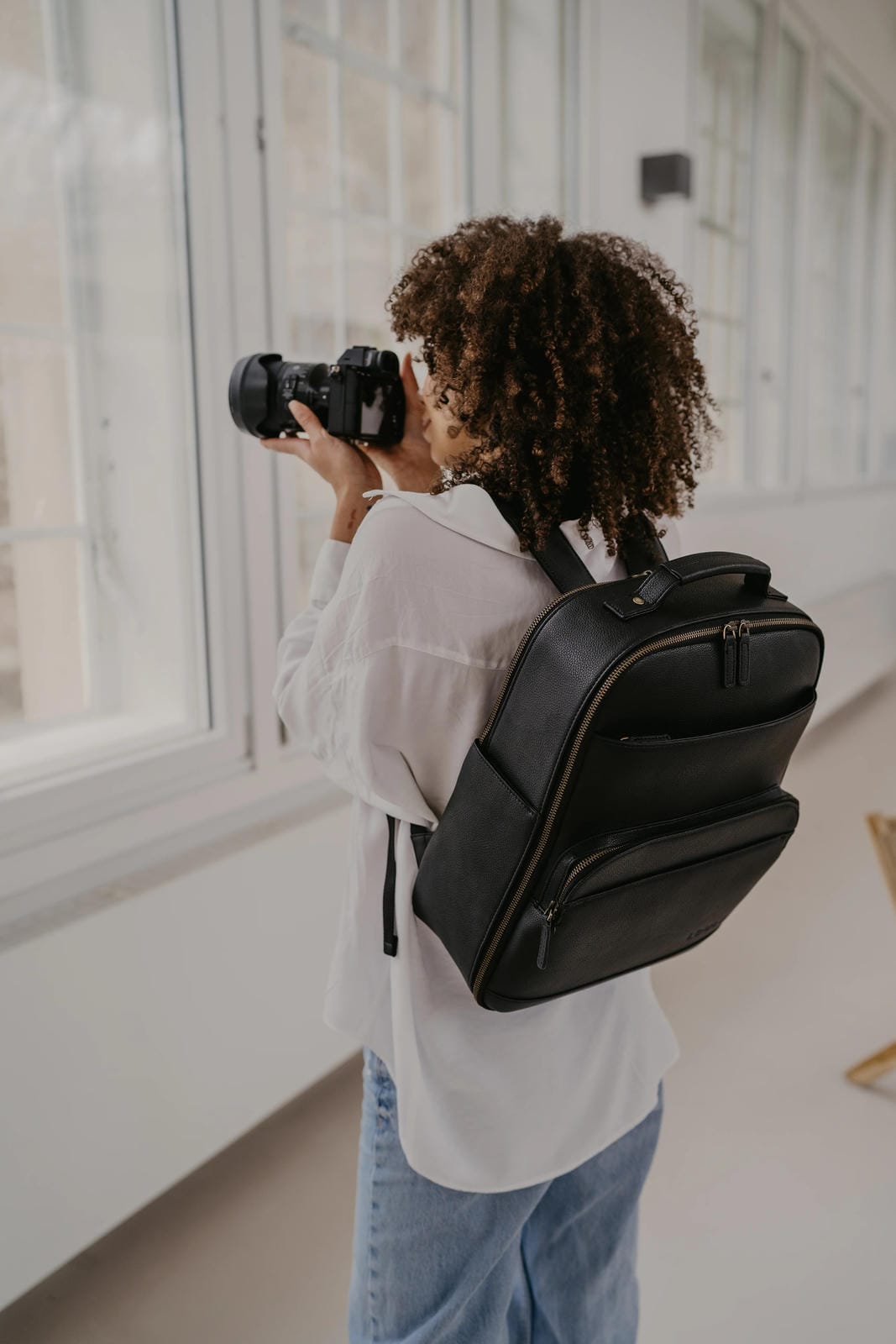 Person with a camera and backpack taking a photo in a room with large windows.