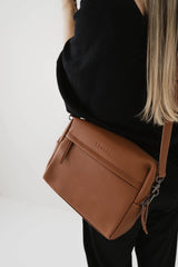 Bolso marrón con el logotipo de la marca visible llevado por una persona sobre un fondo blanco.