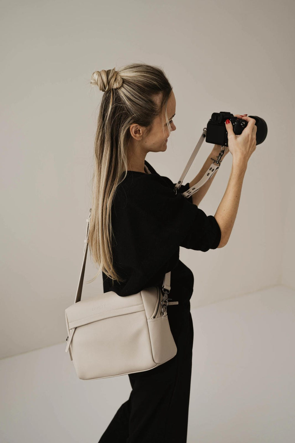 Persona sosteniendo una cámara con un bolso beige sobre un fondo liso.