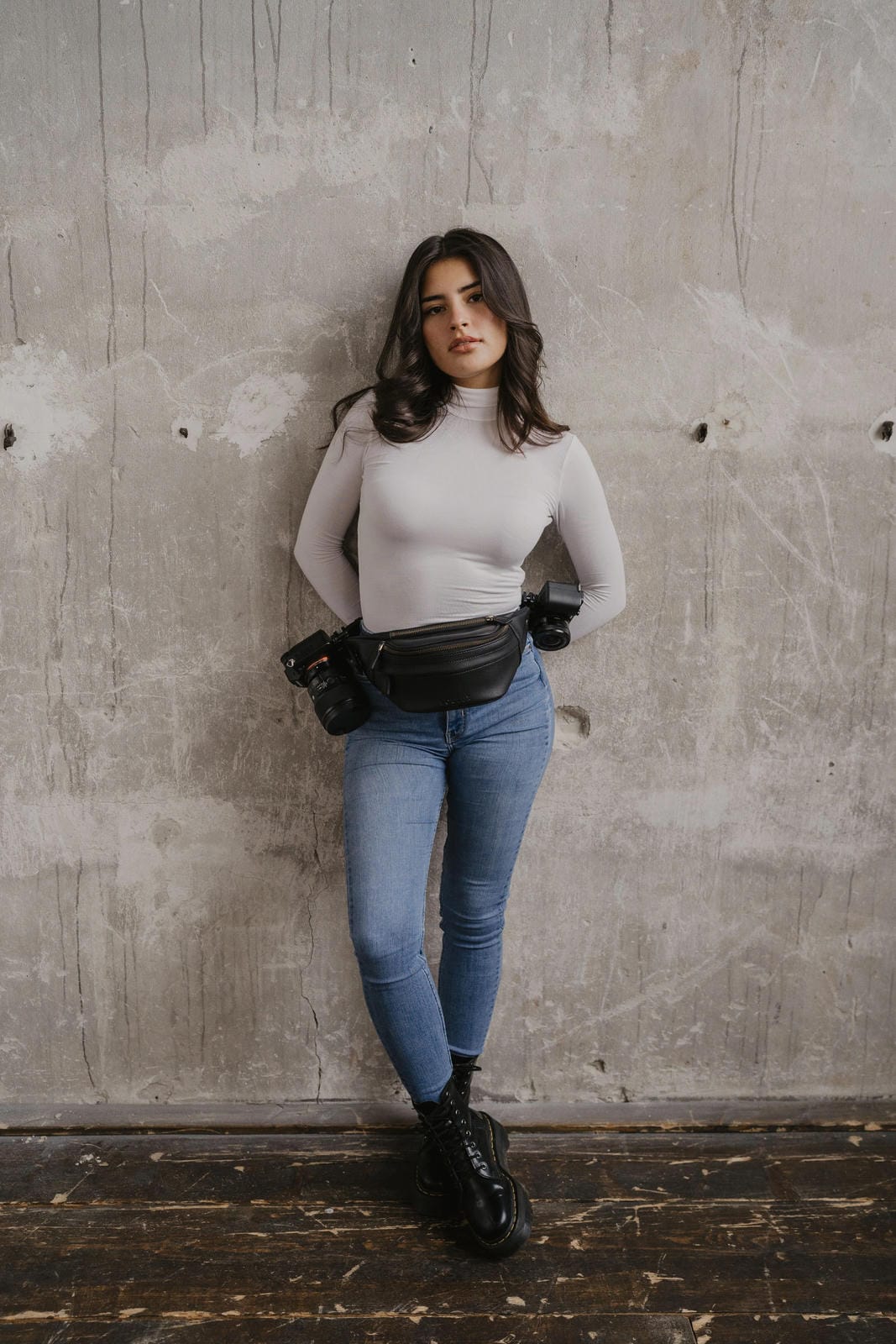 Woman wearing a white turtleneck, blue jeans, and black boots against a concrete wall.