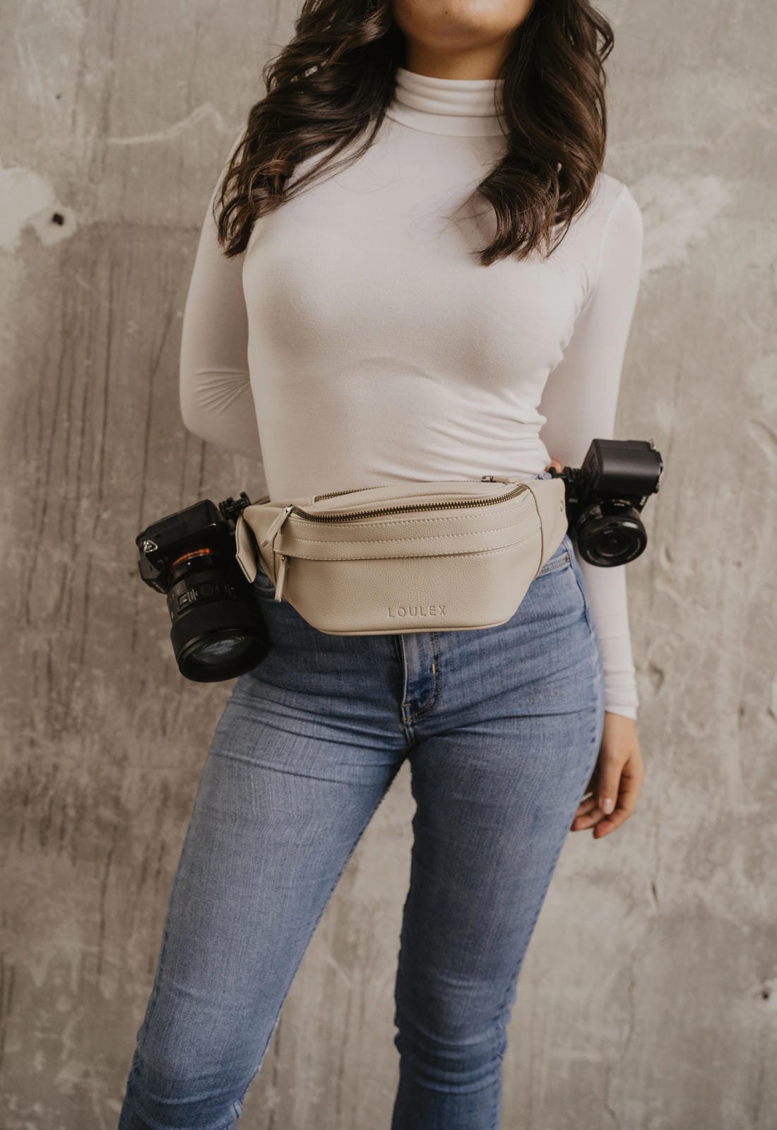 Person wearing a beige waist bag with camera equipment against a concrete wall.