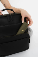 Bolso de cuero negro con una correa verde sostenido por una mano sobre un fondo blanco.