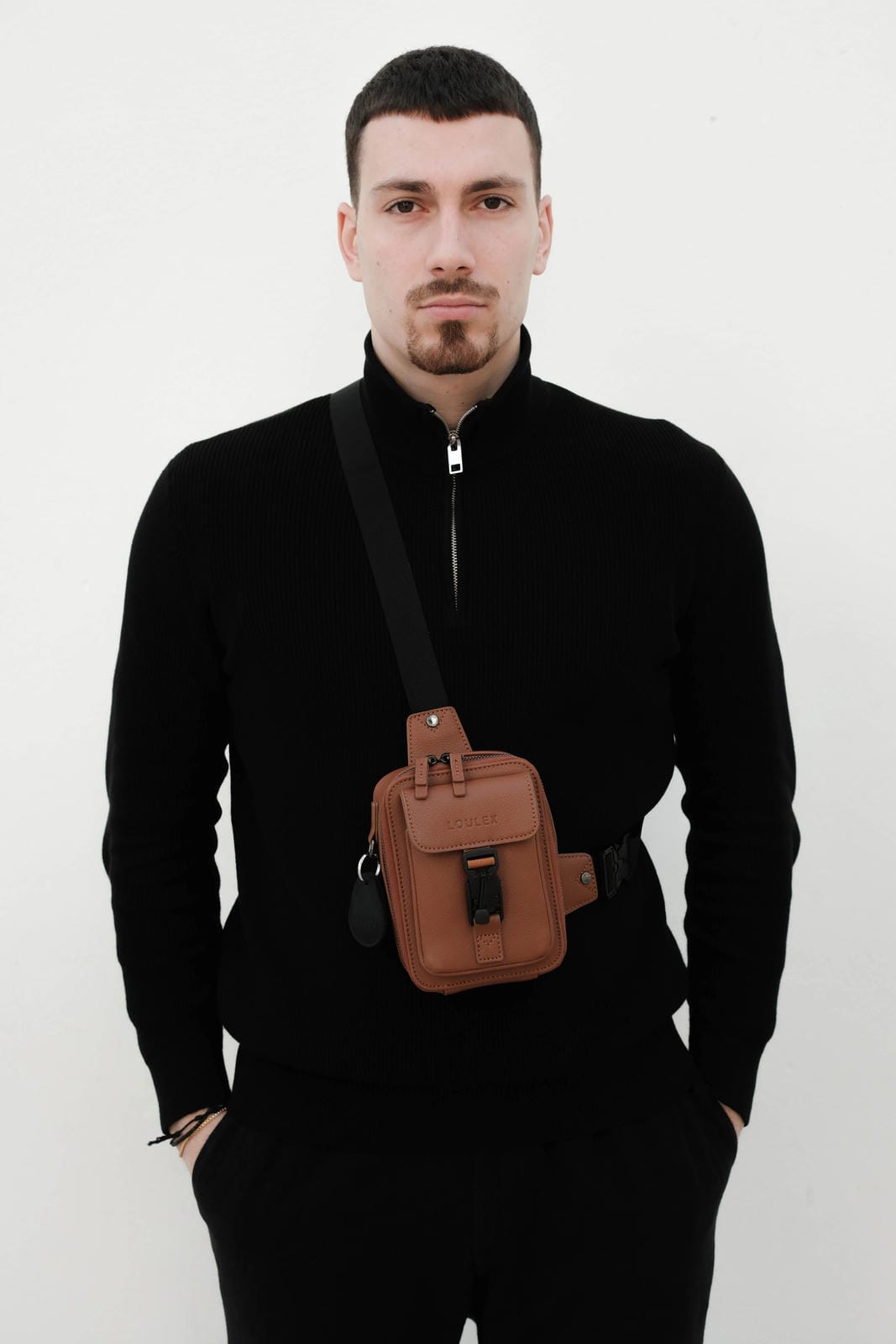 Man wearing a black sweater with a brown leather bag over his shoulder against a white background