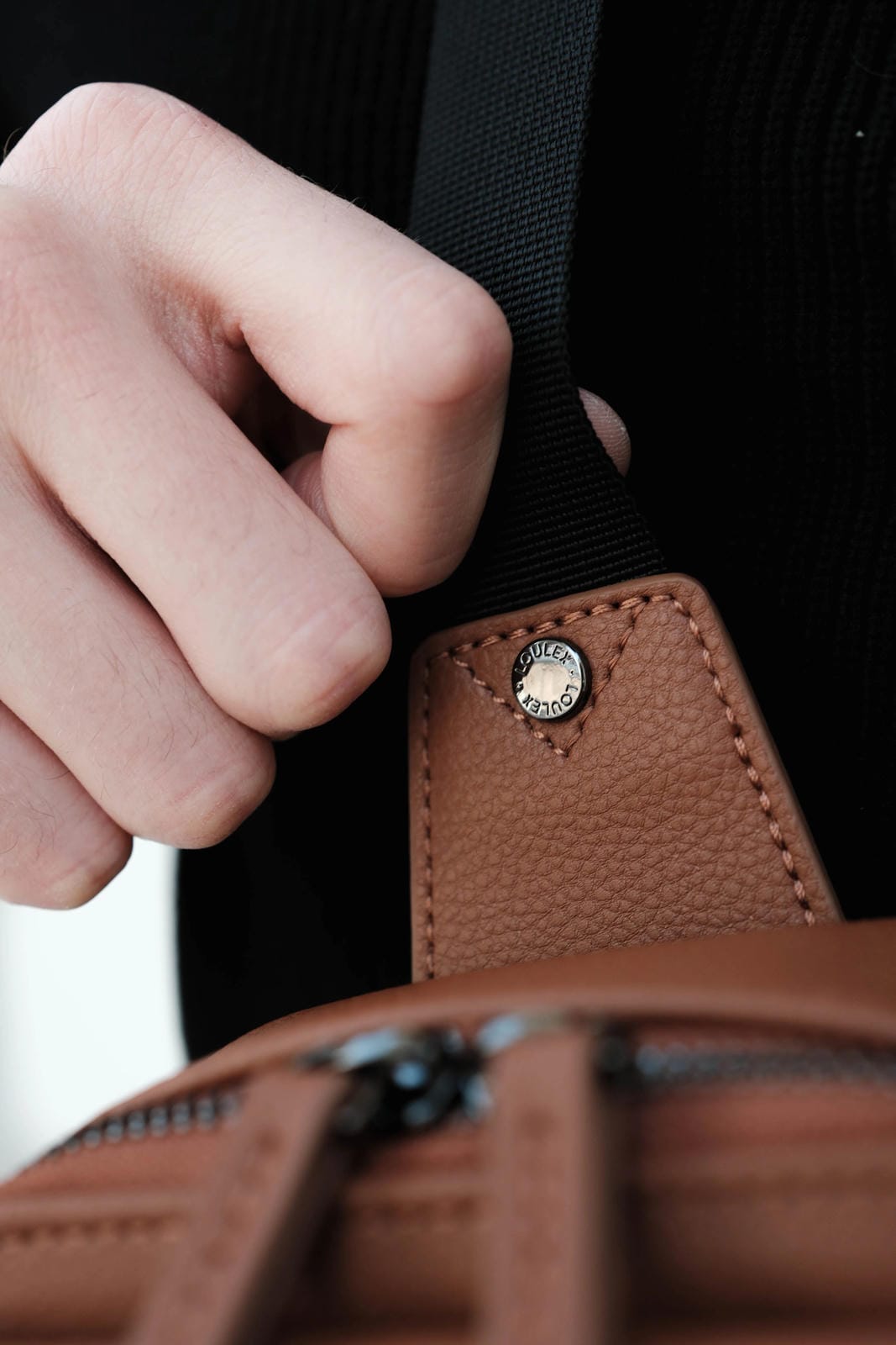 Close-up of a hand adjusting a strap on a brown leather bag with a visible brand logo.