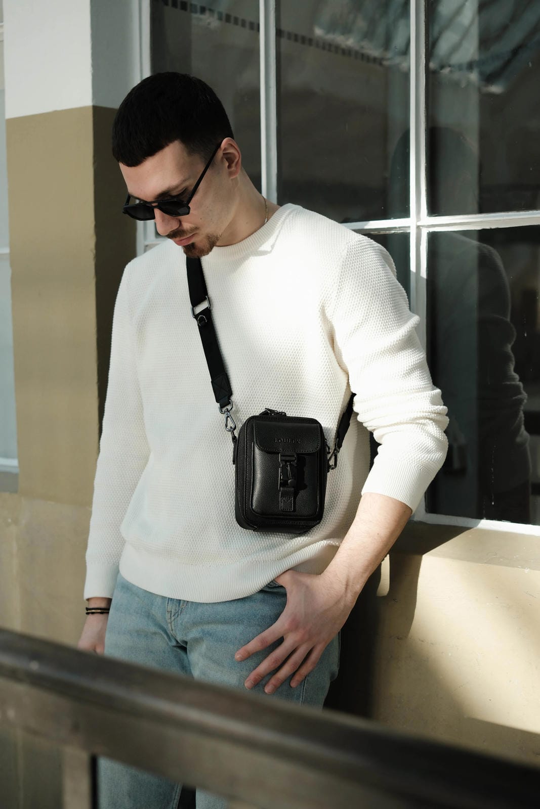 Man wearing a white sweater and sunglasses with a black bag, standing in front of a reflective surface.