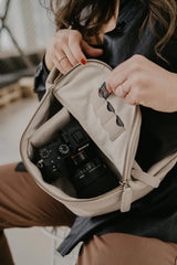 Persona che apre una borsa beige per rivelare una macchina fotografica al suo interno, con sfondo sfocato.