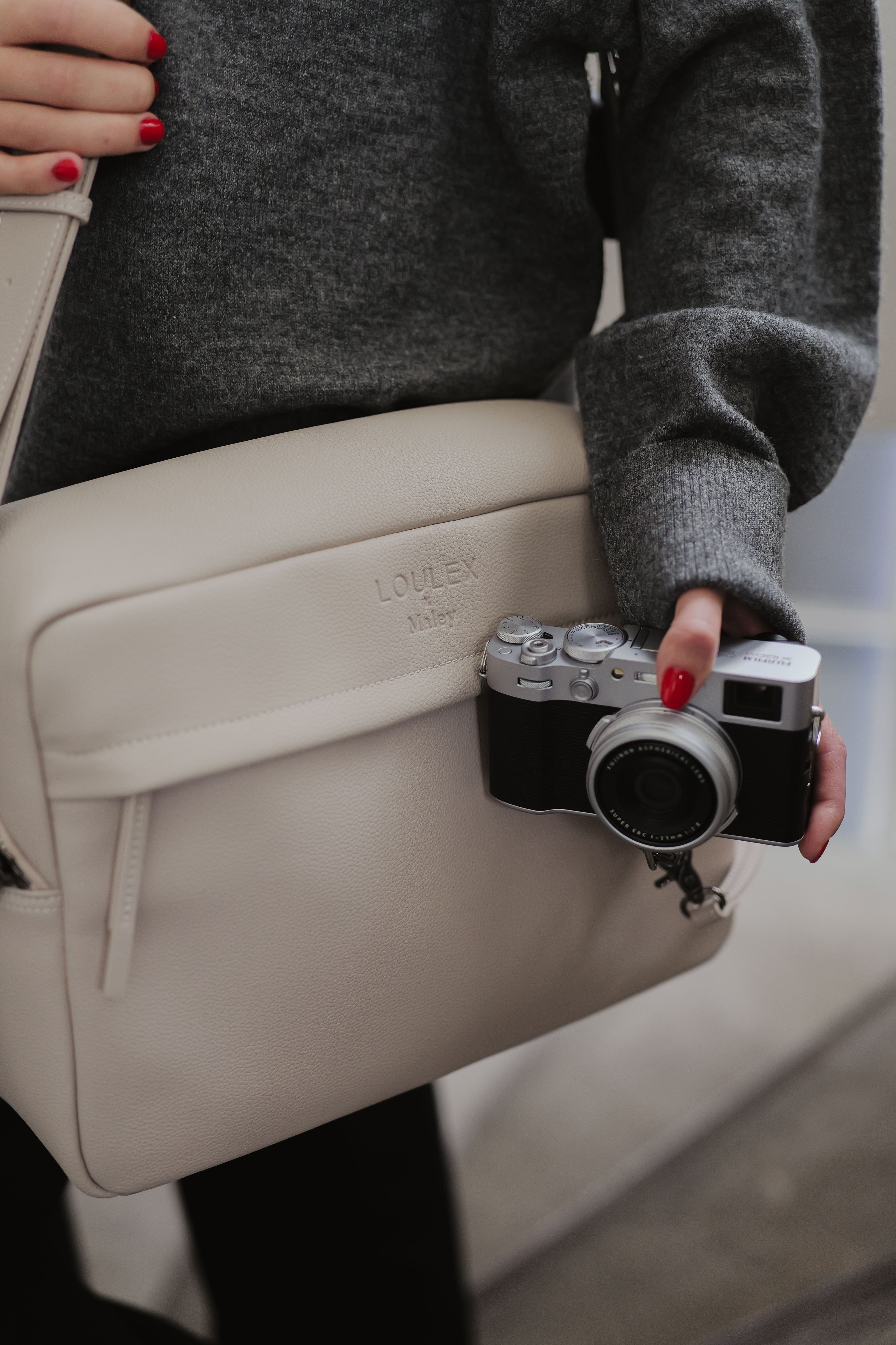 Persoon met een beige tas met een camera eraan, gekleed in een grijze trui.