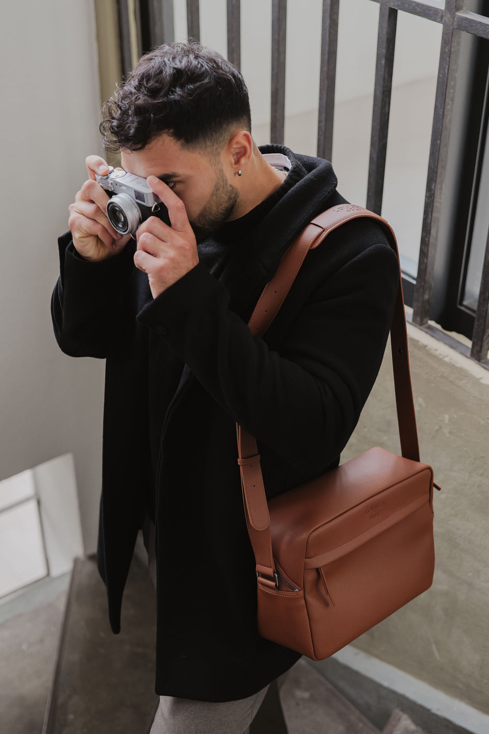 Mężczyzna robiący zdjęcie aparatem fotograficznym, ubrany w czarny płaszcz i niosący brązową skórzaną torbę.