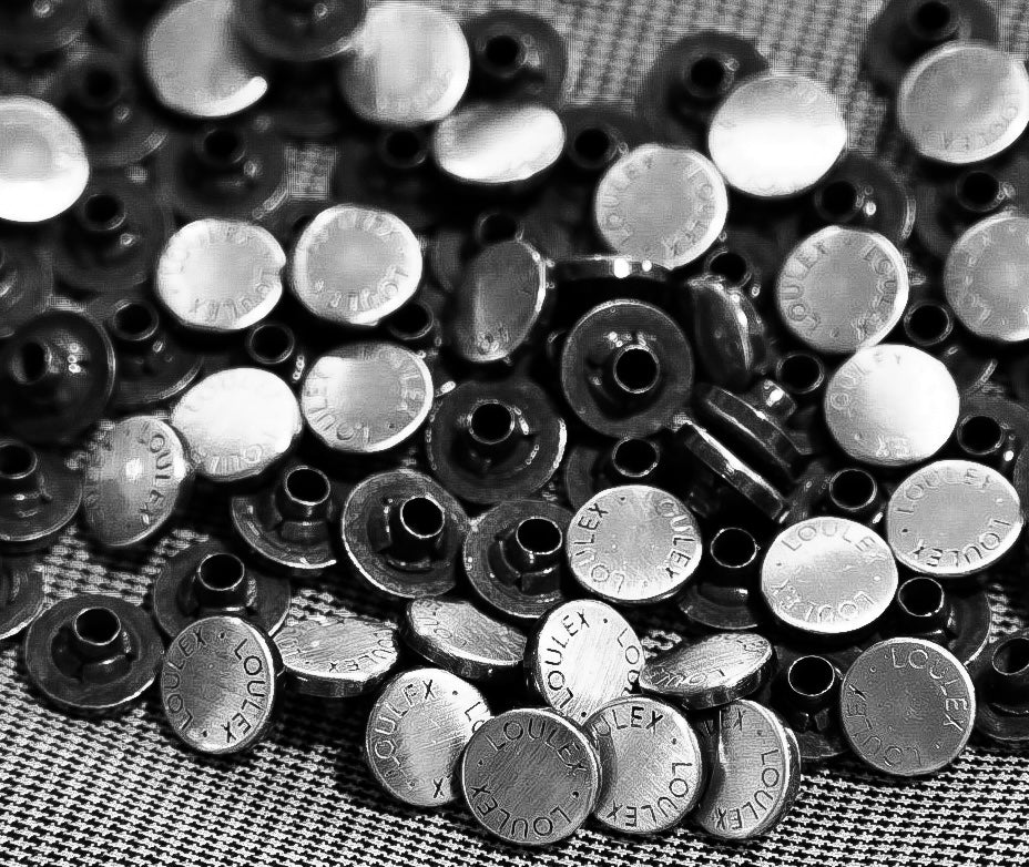 A pile of custom silver-toned metal buttons with the loulex logo, showcasing the durable materials and artisan craftsmanship of luxury photography accessories.