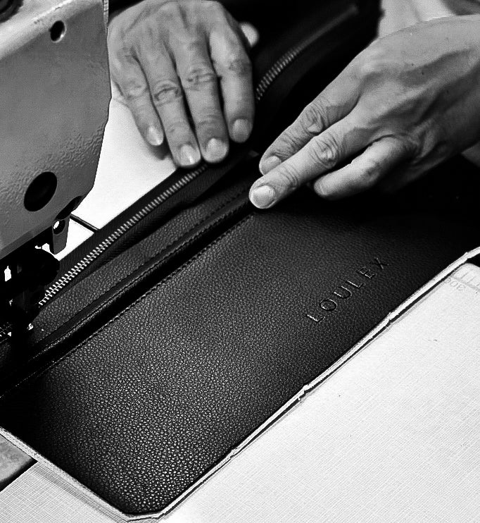 Close-up of an artisan’s hands sewing a black pebbled leather piece with the loulex logo embossed on it.