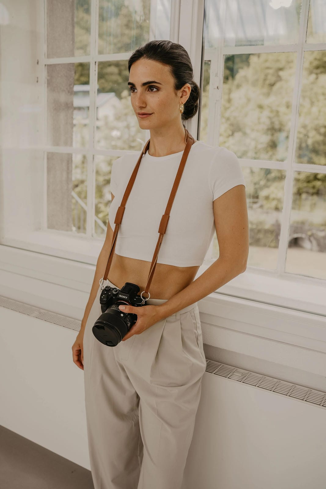 Femme tenant un appareil photo avec une sangle, debout dans une pièce avec de grandes fenêtres.