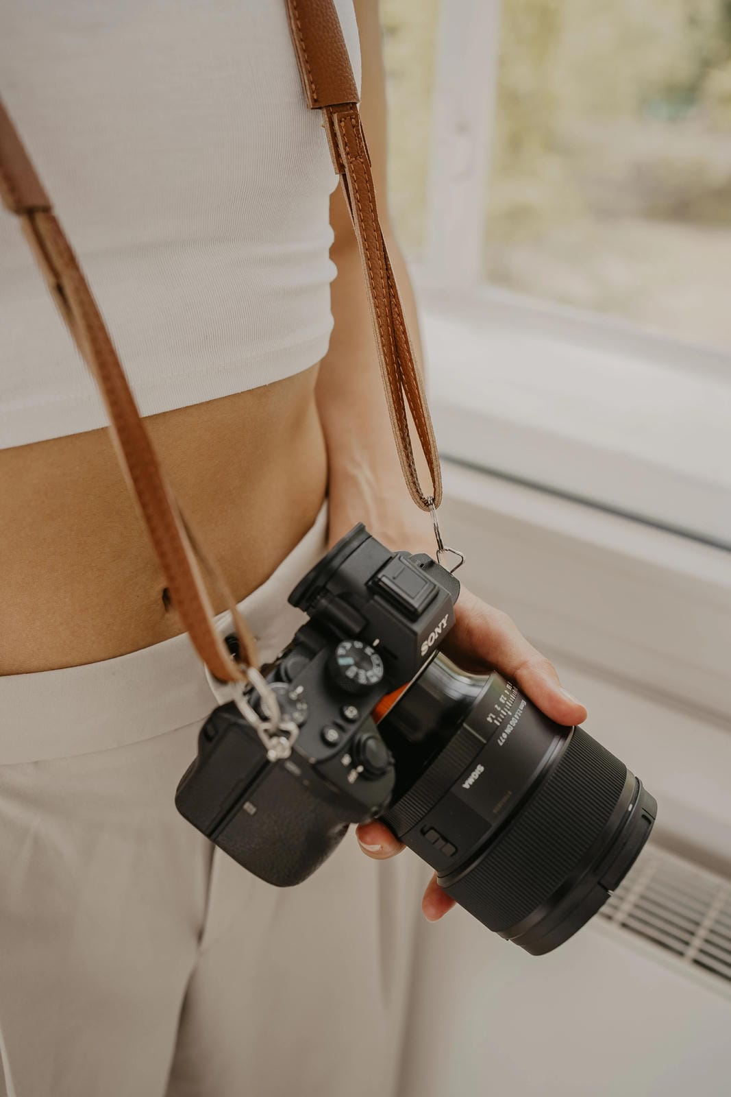 Personne tenant un appareil photo avec une sangle, debout à l'intérieur près d'une fenêtre.