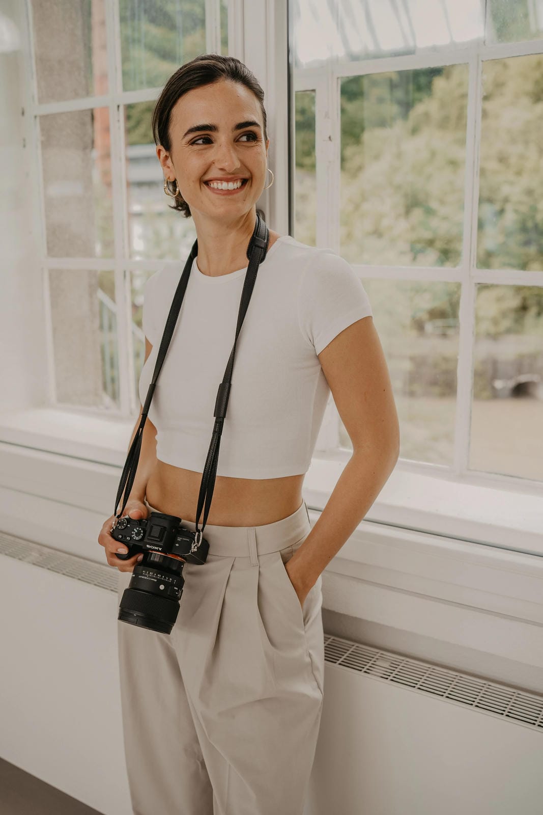 Femme tenant un appareil photo, debout dans une pièce avec de grandes fenêtres.