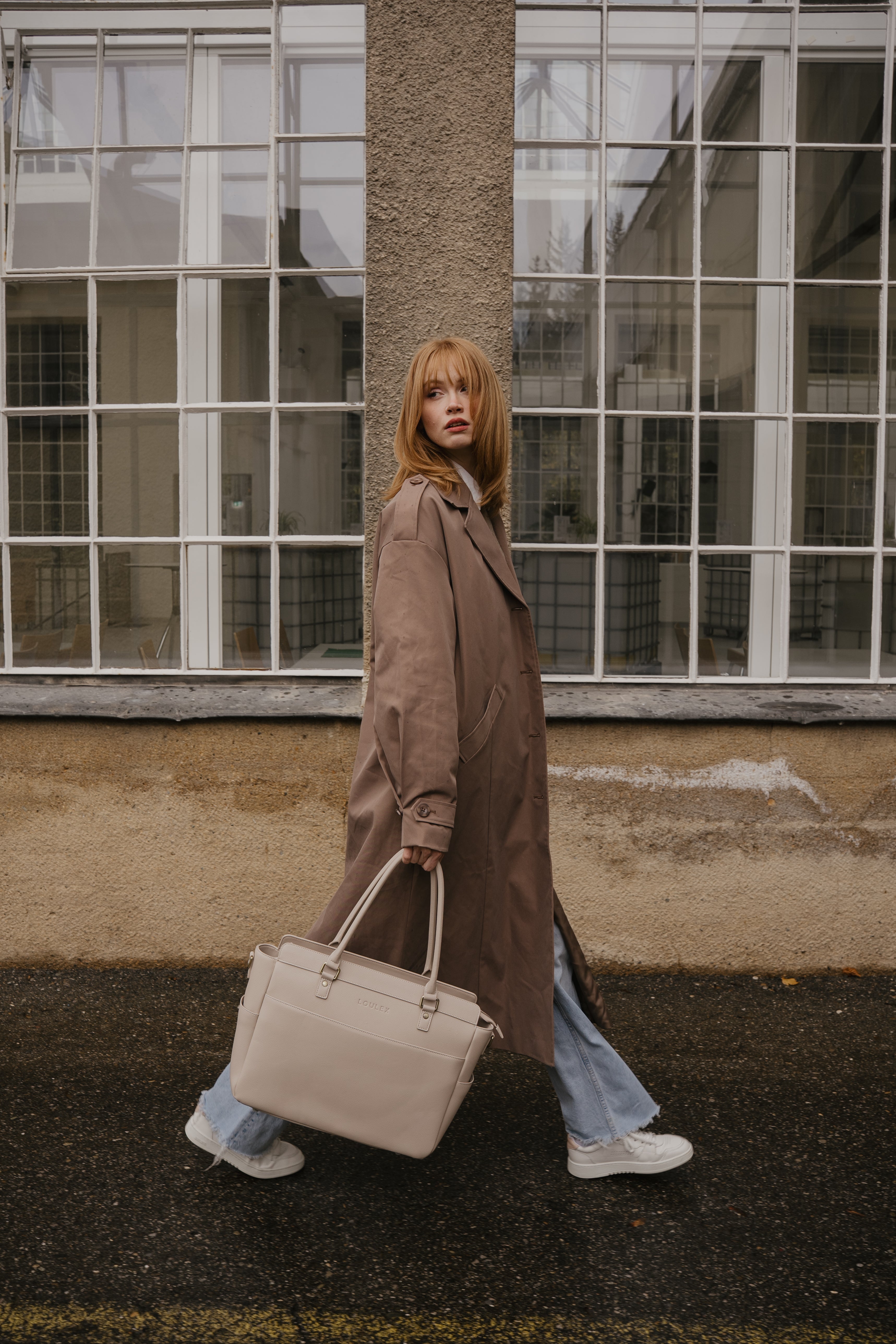 Femme vêtue d'un long manteau et tenant un sac beige, debout devant un bâtiment aux grandes fenêtres.