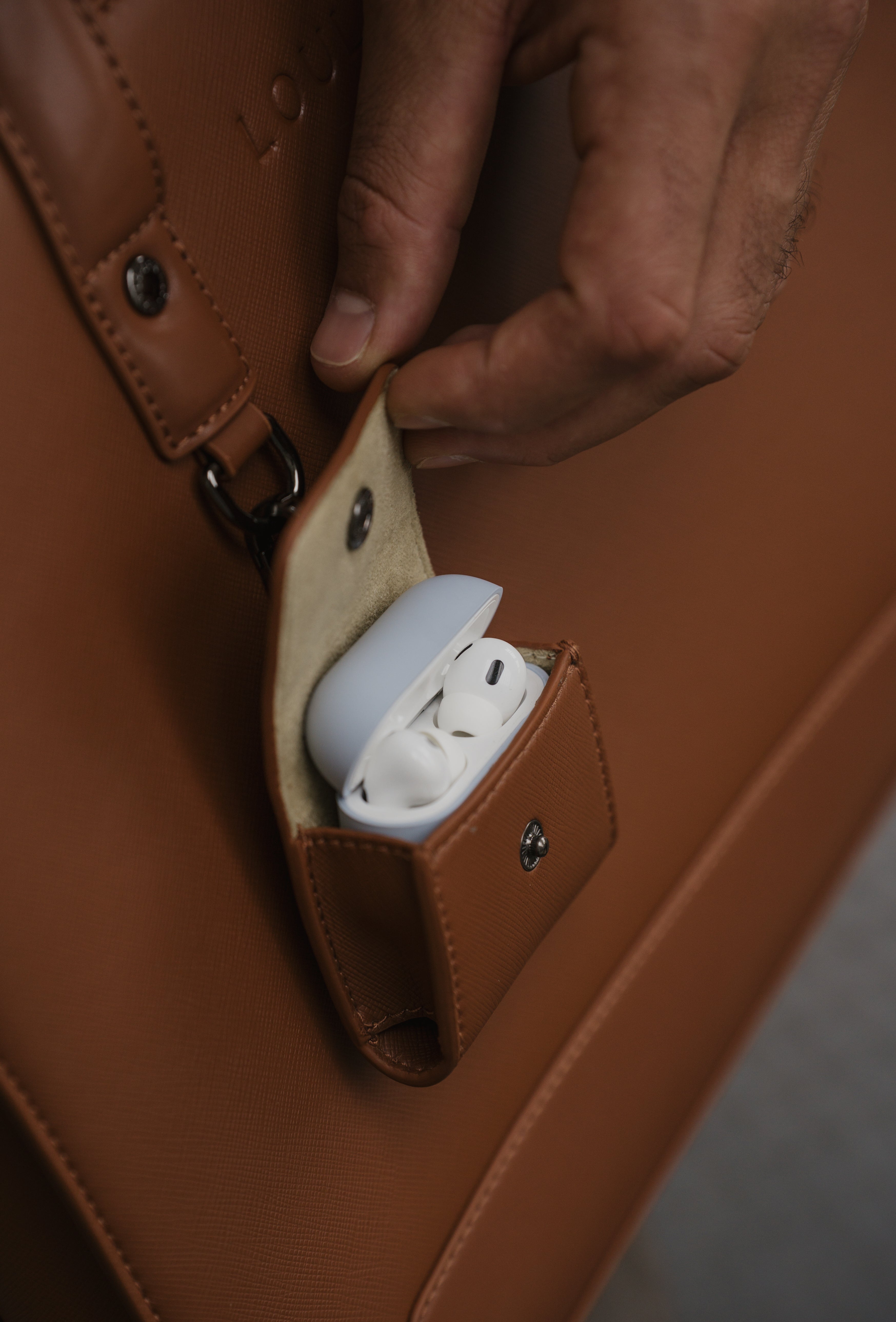 Brown leather case with a small pocket containing white earbuds, held by a hand.