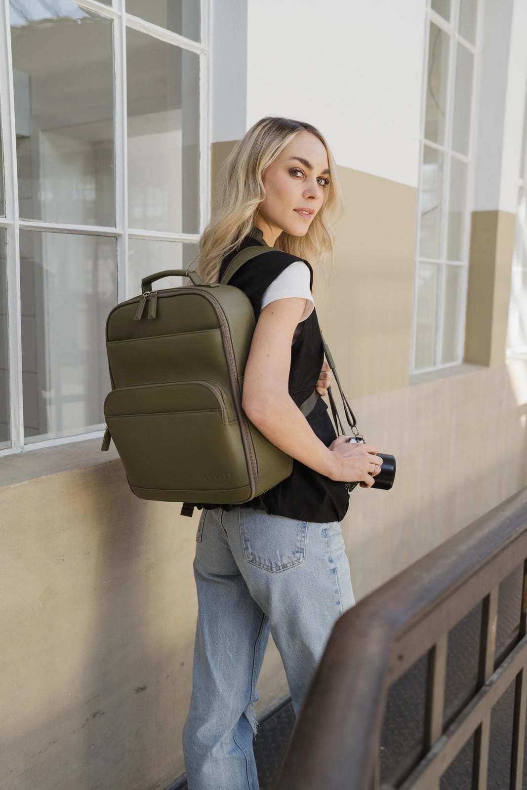 Femme avec un sac à dos vert debout contre un bâtiment.