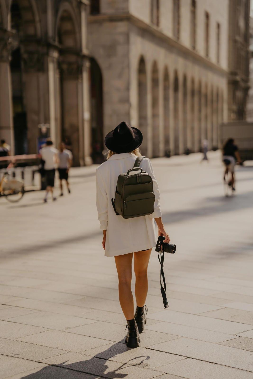 Person walking on a city street with a backpack and camera, wearing a hat.