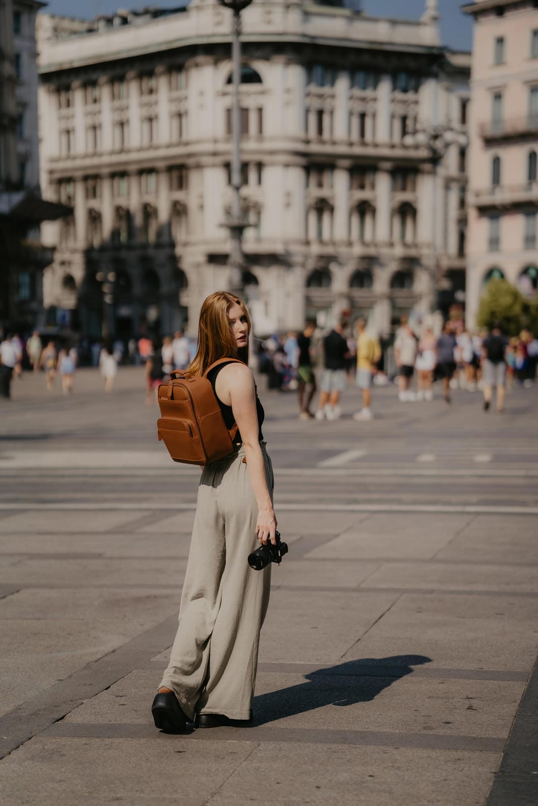 Woman with a brown backpack standing in a city square with people and buildings in the background