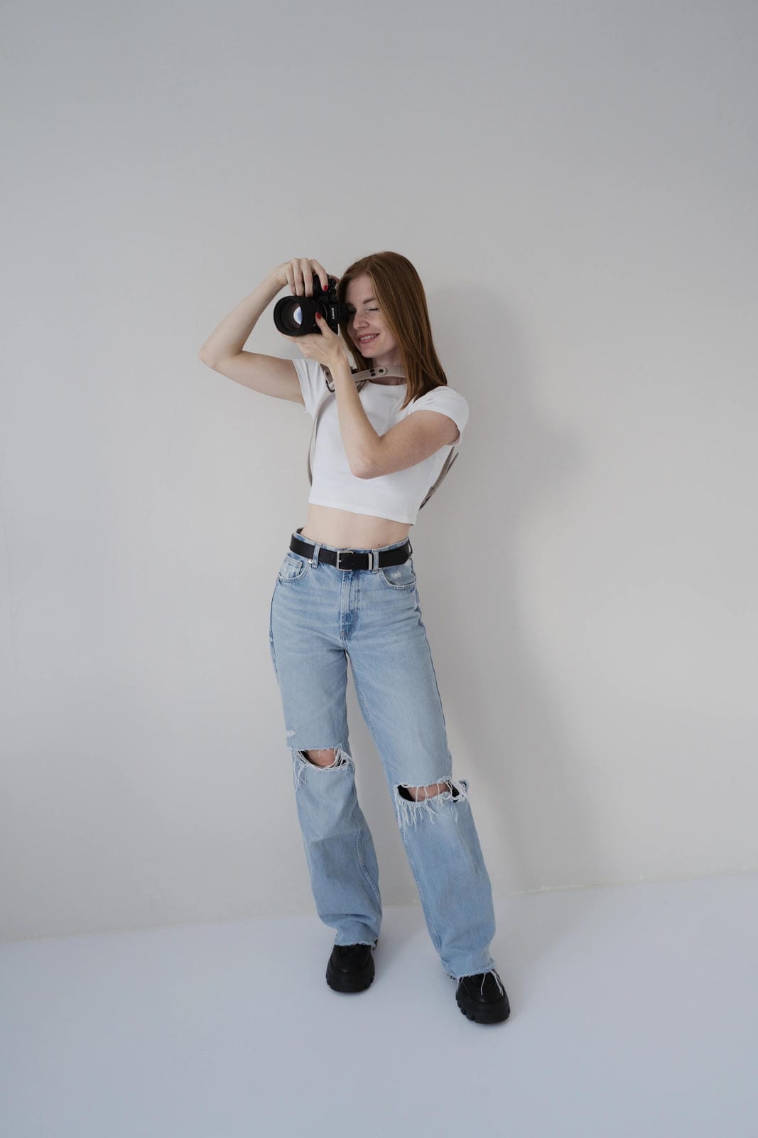 Person wearing a white crop top and light blue jeans, holding a camera against a plain background