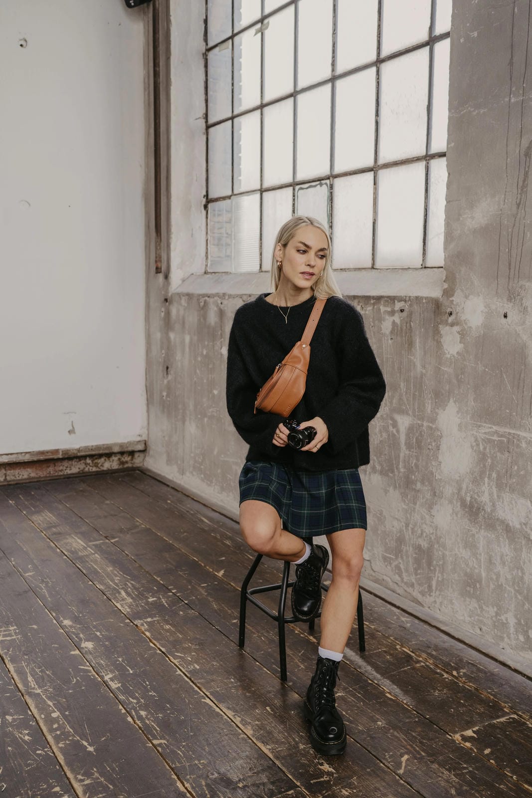 Woman sitting on a stool in an industrial-style room with large windows.