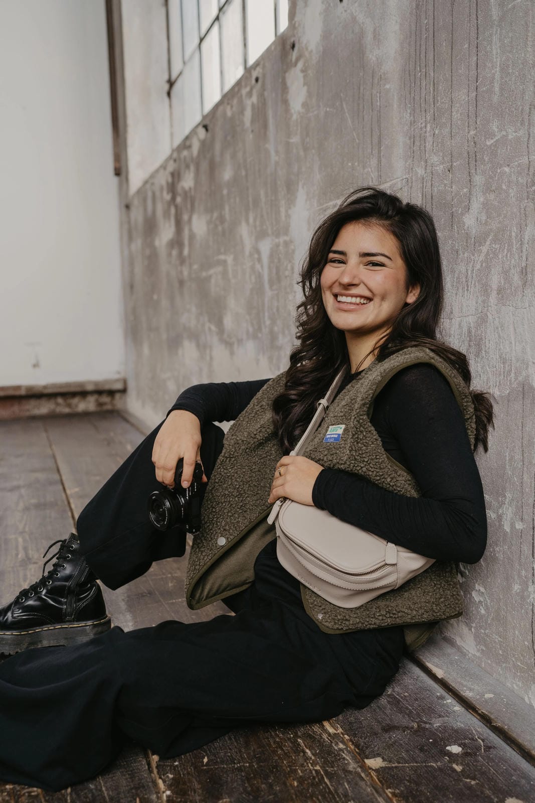 Woman sitting against a wall, wearing a brown vest and black pants.