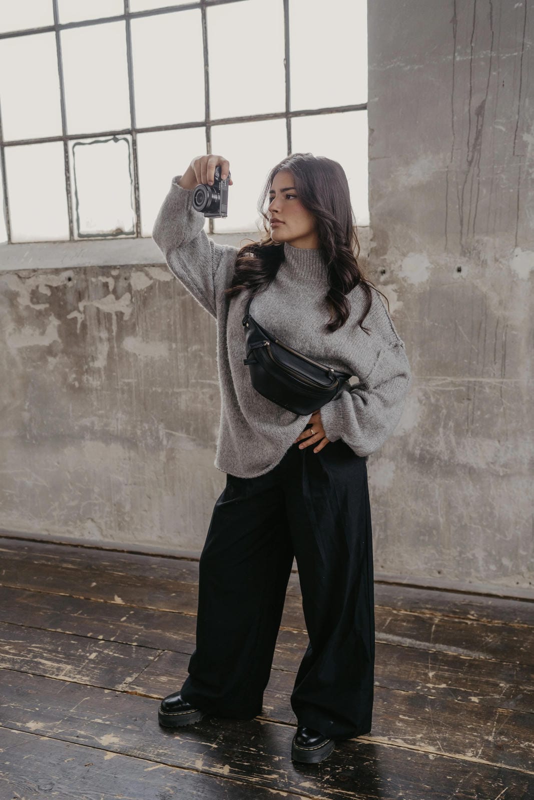 Person taking a selfie in a gray sweater and black pants with a black bag in an industrial setting.
