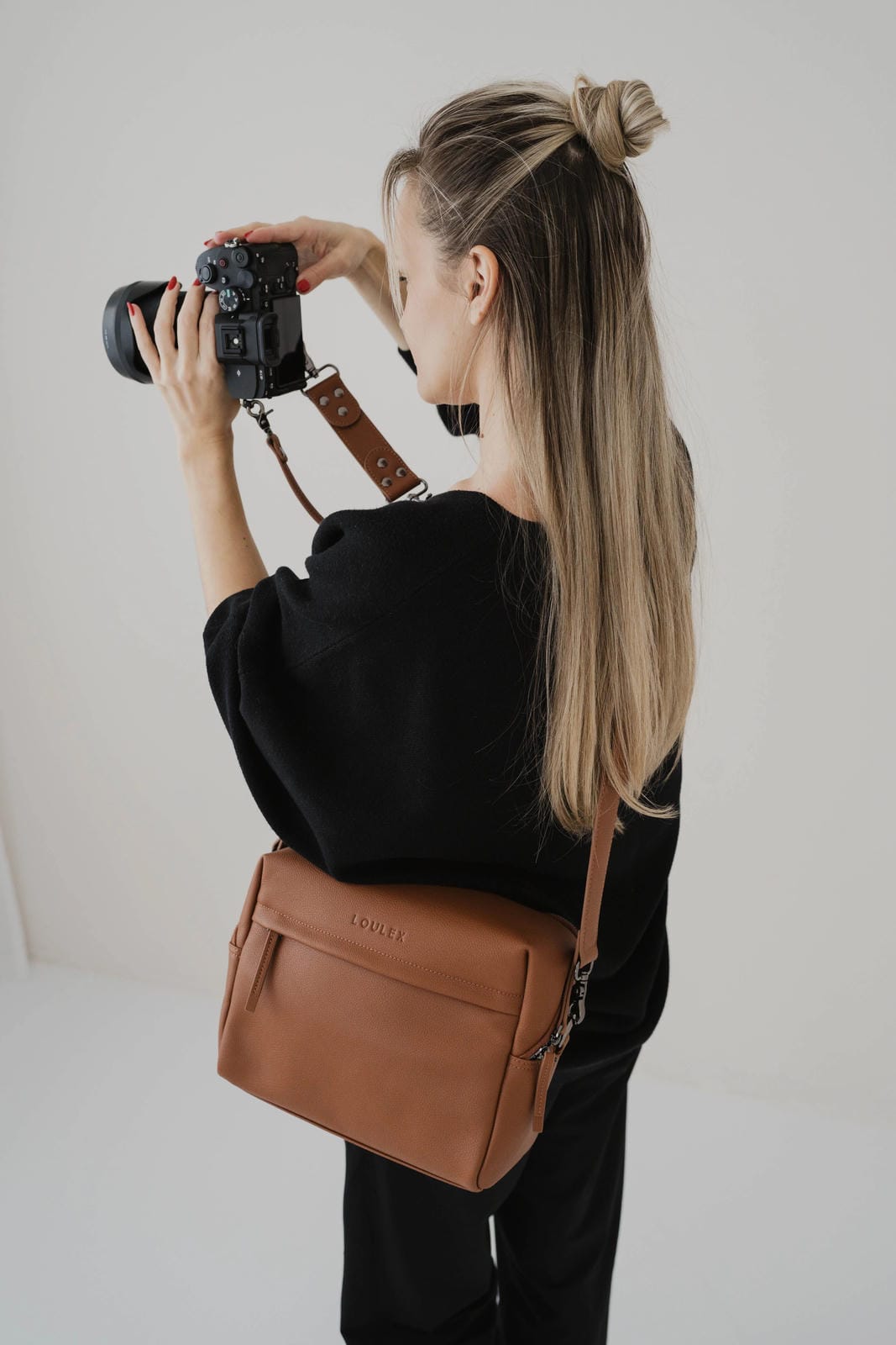 Person holding a camera with a brown leather bag over their shoulder on a plain background