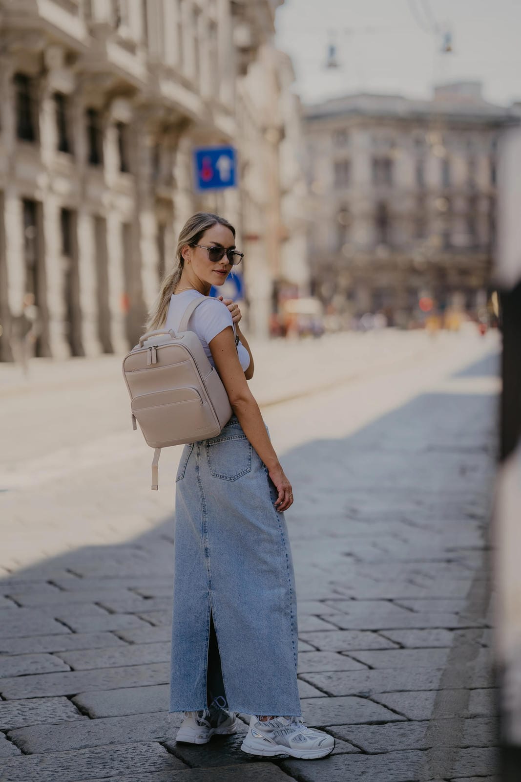 Woman walking on a city street wearing a beige backpack and sunglasses.