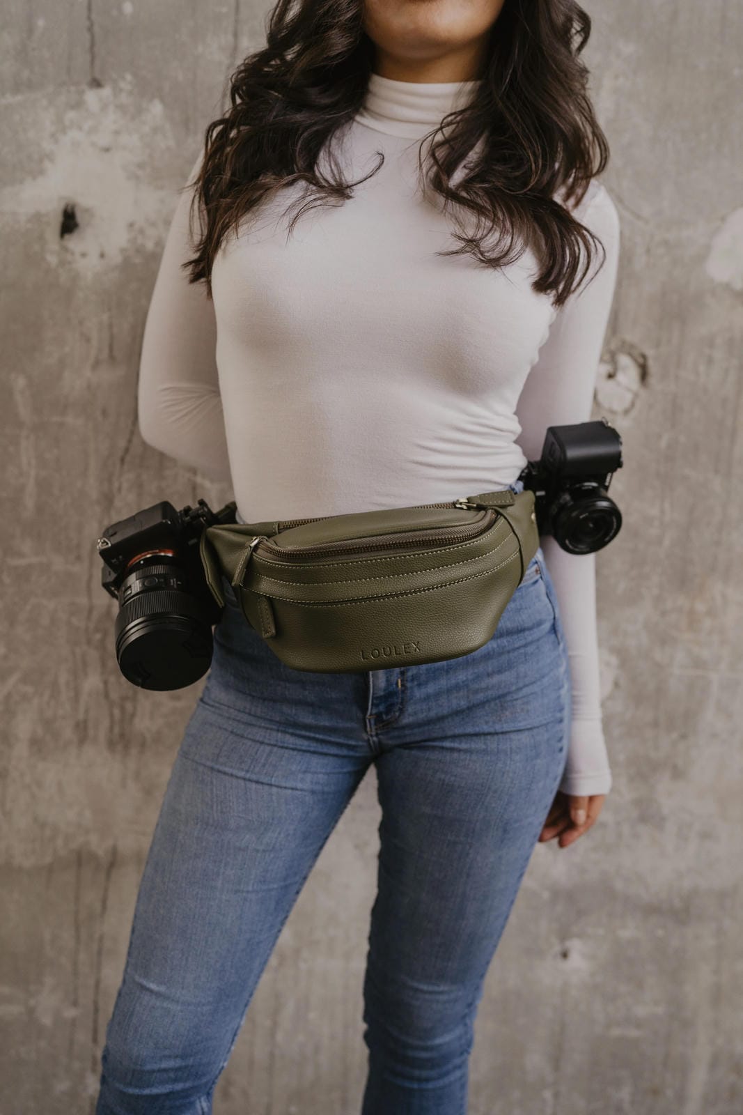 Person wearing a green waist bag with cameras attached against a concrete wall.