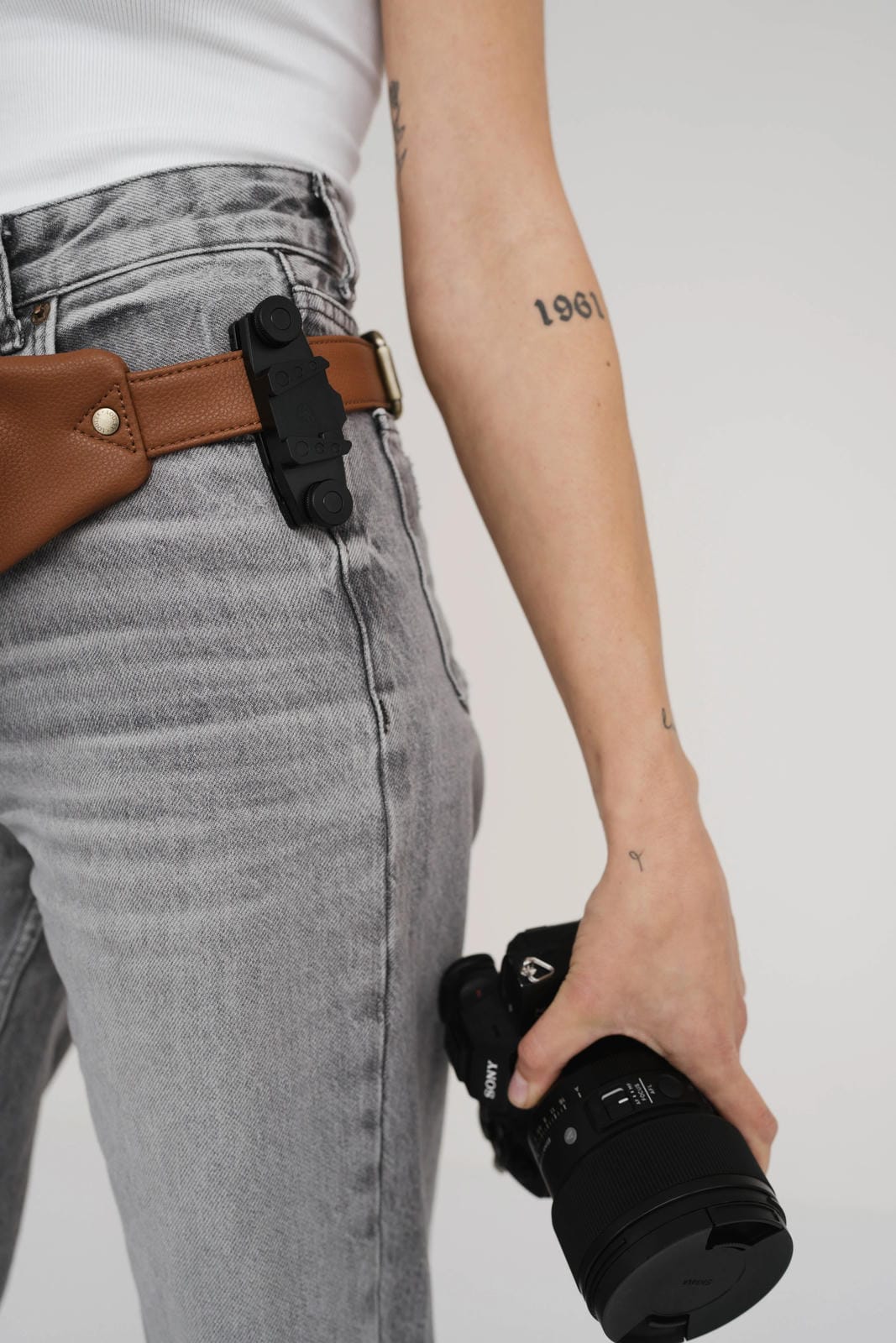 Person wearing a brown belt with a camera attached, holding a black camera lens.