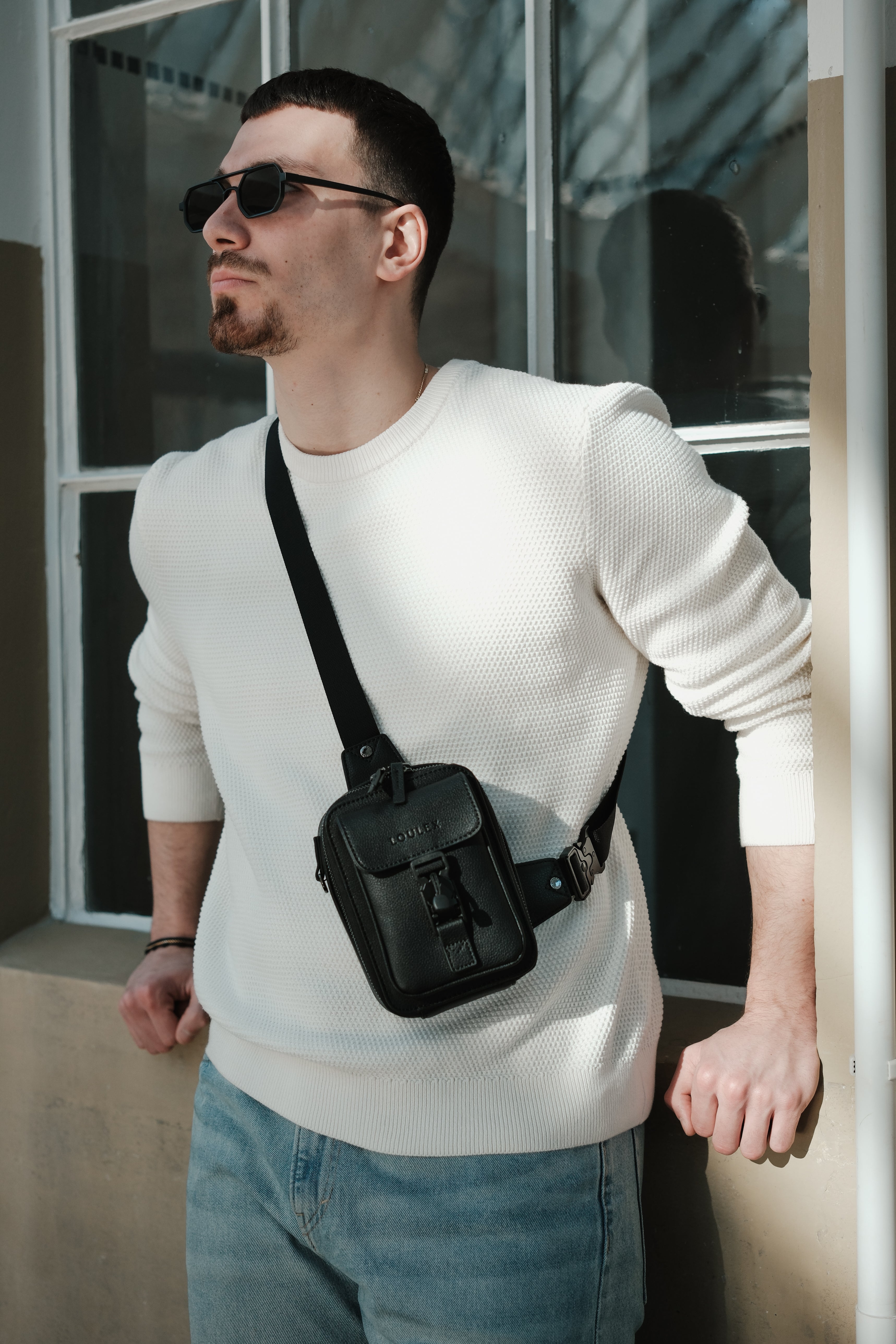 A person wearing sunglasses and a white sweater is carrying a black LOULEX streetbag with a shoulder strap.