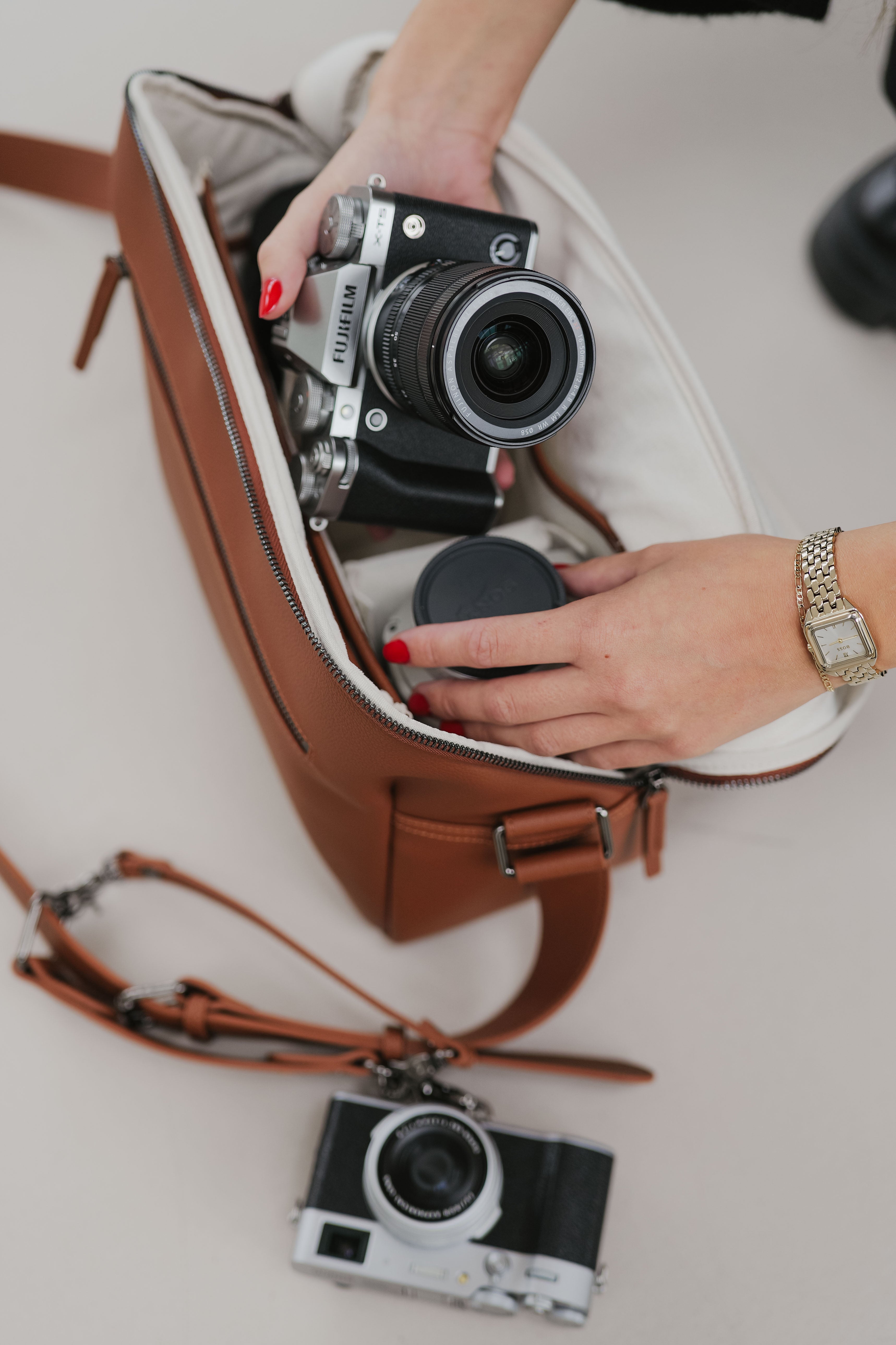 Persoon die een camera in een bruine leren tas organiseert met een andere camera op een wit oppervlak.
