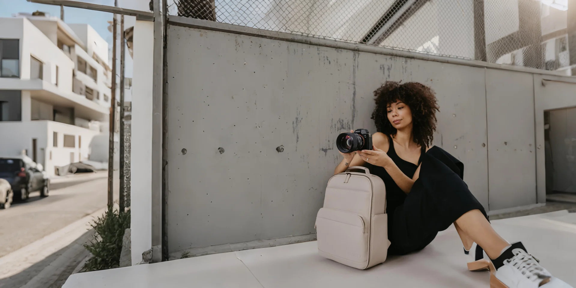 Woman sitting on a ledge with a beige backpack and camera, urban setting
